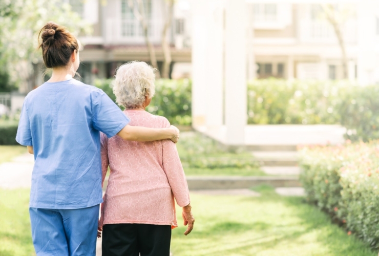 A young woman caregiver in blue scrubs puts her arm around an elderly woman as they walk outside together through a green landscape.