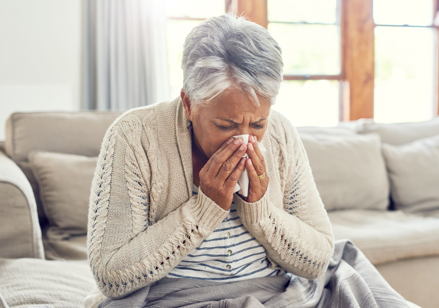 Senior woman, sick during flu season and blowing nose at home from virus, allergy and illness on couch. Tired, tissue and elderly female person in a house lounge with sneeze from covid and flu on a sofa with allergies.