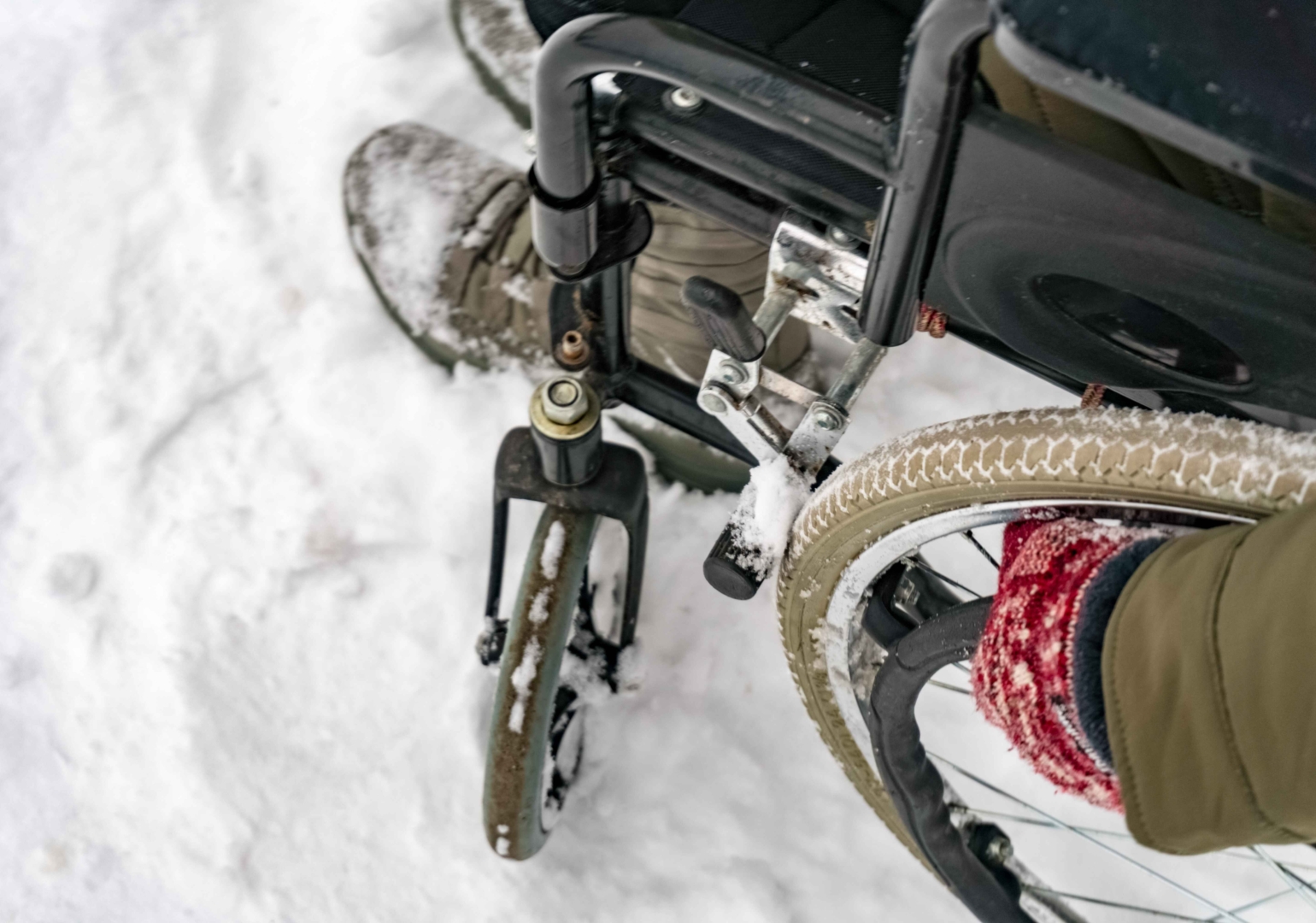 Close-up of a person in a wheelchair navigating through snow. The wheelchair's wheels and the person's booted feet are partially covered in snow, indicating cold weather conditions. The person is also wearing red knit gloves, suggesting winter clothing.