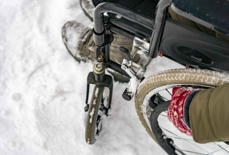 Close-up of a person in a wheelchair navigating through snow. The wheelchair's wheels and the person's booted feet are partially covered in snow, indicating cold weather conditions. The person is also wearing red knit gloves, suggesting winter clothing.