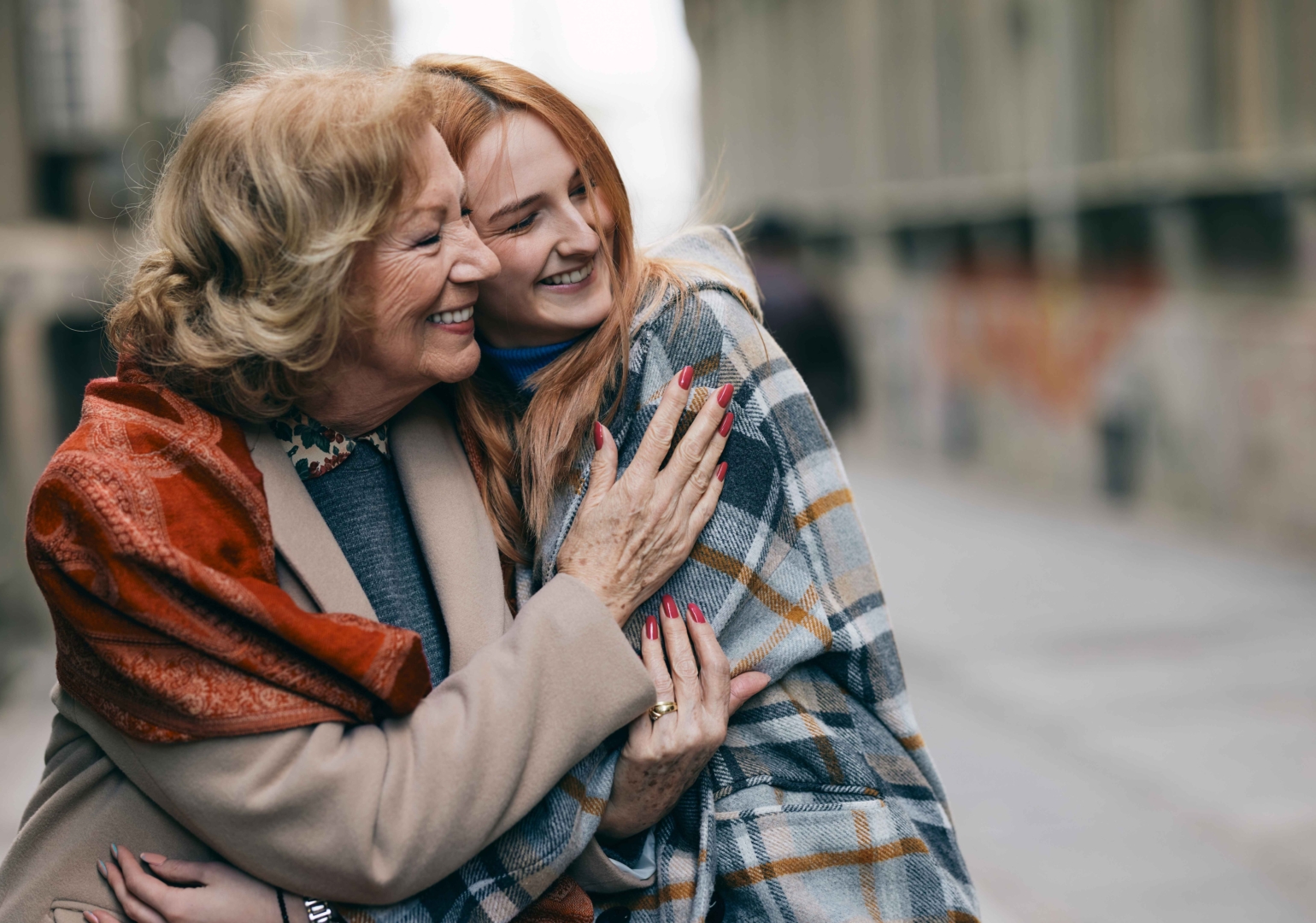 An elderly woman and a younger woman, wrapped in cozy shawls, embrace and smile warmly while outdoors on a chilly day. The image captures a joyful moment of connection and affection.