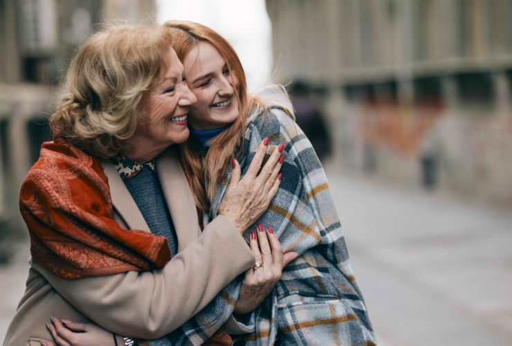 An elderly woman and a younger woman, wrapped in cozy shawls, embrace and smile warmly while outdoors on a chilly day. The image captures a joyful moment of connection and affection.