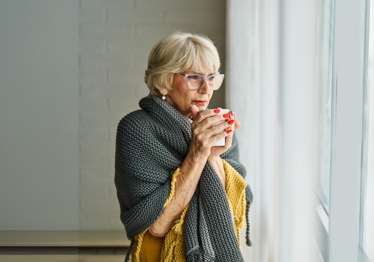 An elderly woman wrapped in a cozy gray and yellow knit blanket holds a warm mug while looking out a window, enjoying a peaceful winter moment indoors.