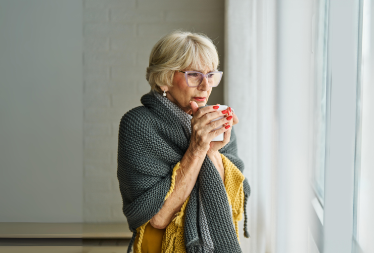 An elderly woman wrapped in a cozy gray and yellow knit blanket holds a warm mug while looking out a window, enjoying a peaceful winter moment indoors.